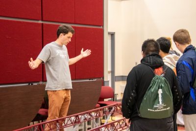 A Virginia Tech student raises his hands wide apart over top of a red metal span. A group of high schoolers stand on the other side watching.