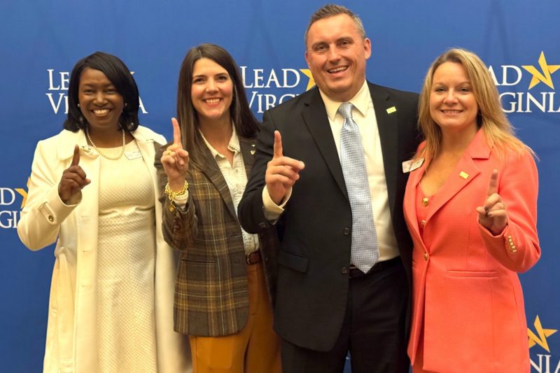 Four people pose for a photo holding up their pointer fingers in front of a blue Lead Virginia backdrop.