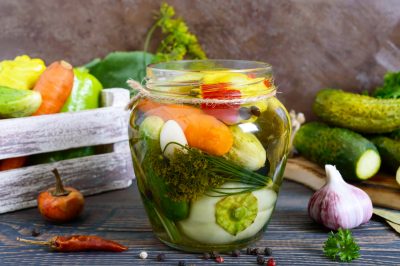 A jar of pickled vegetables, including carrots, cucumbers, and garlic, sits on a wooden surface surrounded by fresh cucumbers, garlic, and peppers. A white wooden crate filled with more fresh vegetables is in the background.