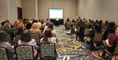 An audience of about 50 people sit in chairs with their backs to the camera in a conference room with plain beige walls and a loud-patterned carpet. A man in a suit stands and speaks next to a screen at the front of the room.