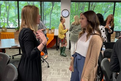 Two women talk in a roomful of other women.